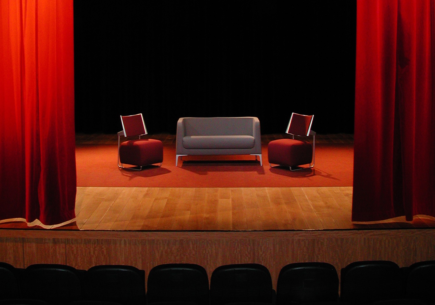 Scène de spectacle avec rideaux rouges, canapé et fauteuils, face aux sièges de la salle